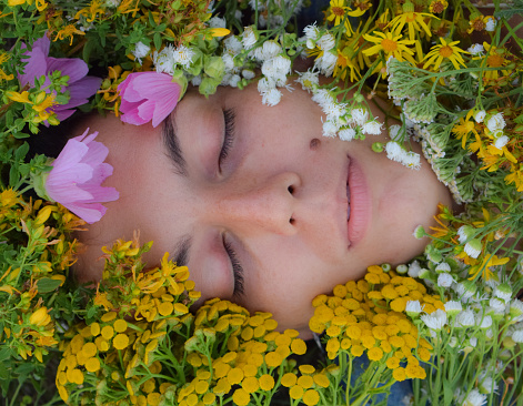 Close-up portrait of a multiracial young woman with closed eyes. Her face adorned by a vibrant array of wildflowers and herbs from the picturesque meadow