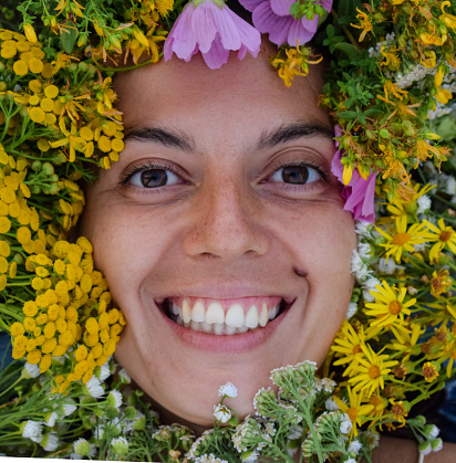 Close-up portrait of a smiling multiracial young woman looking at camera. Her face adorned by a vibrant array of wildflowers and herbs from the picturesque meadow