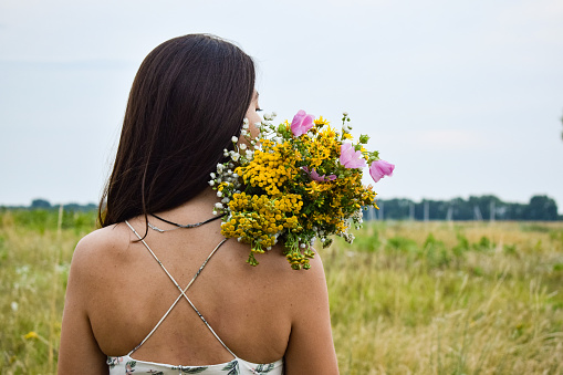 Rear view multiracial young woman holds a bouquet of wildflowers in a picturesque meadow during twilight. The serene scene captures her connection with nature's beauty and the tranquil moments of dusk