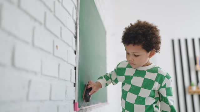 African american cute little child boy wiping clean or erase chalk on green school board. A boy cleaning the chalkboard in classroom at school. Back to School. Education concept