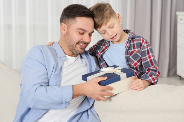 istock Cute little boy presenting his father with gift on sofa at home 1632024268