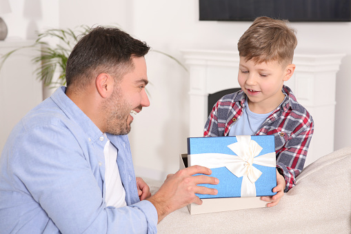 Cute little boy presenting his father with gift on sofa at home