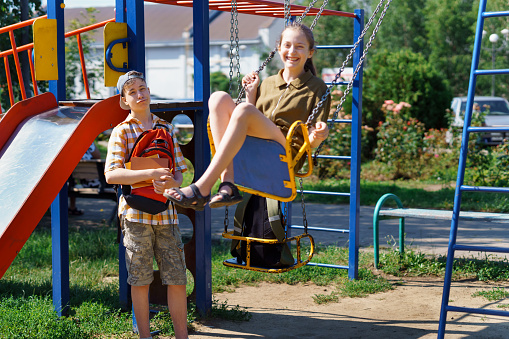 schoolchildren, a teenage boy and a girl play on the playground in the city park after school, a girl rides on a swing, a bright summer day