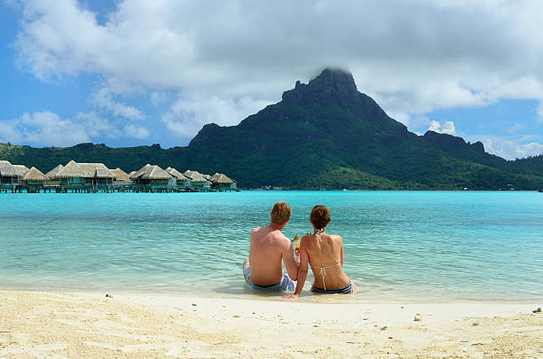 Romantic honeymoon couple near Tahiti A honeymoon couple sharing a cocktail on the sand beach of a luxury vacation resort in the lagoon with a view on the tropical island of Bora Bora, near Tahiti, in French Polynesia. bora bora mountain stock pictures, royalty-free photos & images