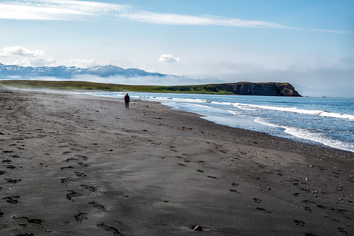 A single woman walks alone on a black sand beach in Tjorneshreppur, Iceland.