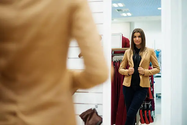 Photo of Beautiful young woman trying on jacket in front of mirror