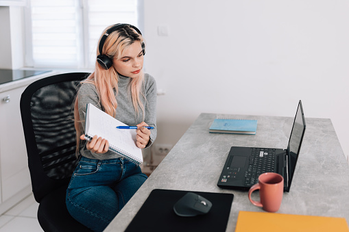 Beautiful responsible young Caucasian woman in home office working from home using laptop and headphones