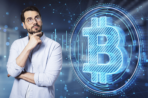 Attractive thoughtful young european businessman with glowing blue polygonal bitcoin hologram on blurry office interior background. Cryptocurrency and financce concept. Double exposure