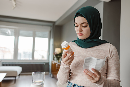 A young Asian woman in a hijab is reading the ingredients on a bottle of pills, while sitting in her living room.