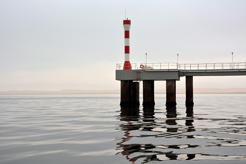 A small lighthouse is located on a sea iron pier above the water surface. The weather is not sunny and cold