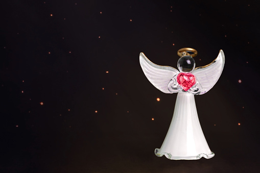 Angel glass figurine holding a red heart on black starry night background with copy space. Spun glass ornament.