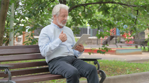 istock Senior Old Man Doing Video Chat on Tablet while Sitting Outdoor on a Bench 1632273026