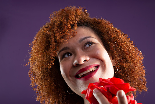 Close-up portrait of a beautiful woman with red curly hair holding a red flower close to her face. Isolated on lilac colored background.