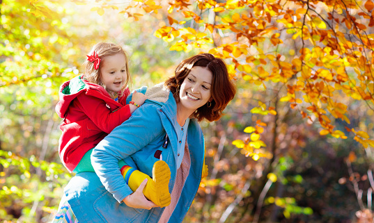 Mother and child play in autumn park. Mom and kid walk in the forest on a sunny fall day. Children playing outdoors with yellow maple leaf. Toddler and parent collect golden leaves. Mom hugging kid.