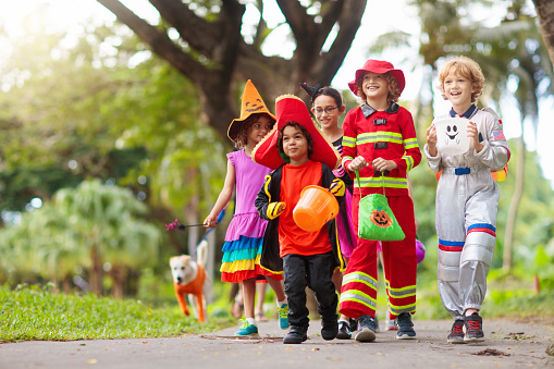 Child in Halloween costume. Mixed race kids and parents trick or treat on street. Little boy and girl with pumpkin lantern and candy bucket. Baby in witch hat. Autumn holiday fun.