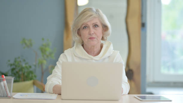 istock Senior Old Woman Working on Laptop Looking toward Camera 1632349919