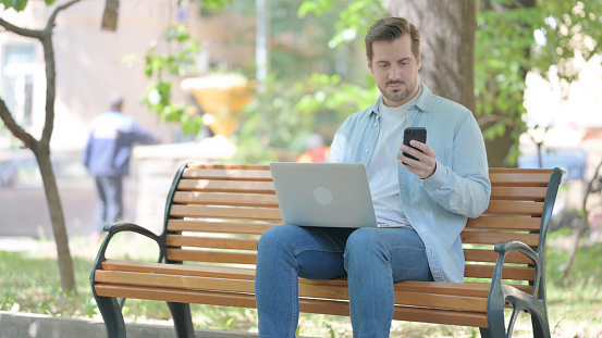 Young Man Using Smartphone and Laptop while Sitting Outdoor