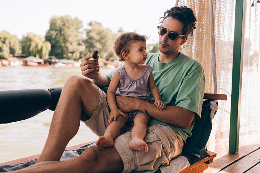 A wide angle view of a Caucasian male with sunglasses holding some sweets while enjoying spending time with his adorable daughter near the river.