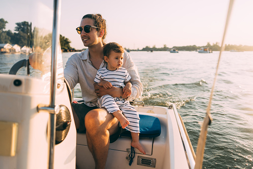 A smiling Caucasian man with sunglasses looking away while holding his adorable daughter. He is on a river boat enjoying his summer vacation.