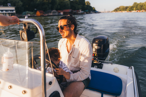 A smiling Caucasian male with sunglasses holding his adorable child while being on a river boat enjoying his summer vacation.