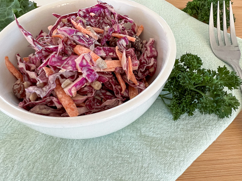 Red cabbage coleslaw salad in a white ceramic bowl, on a green fabric napkin and wooden background