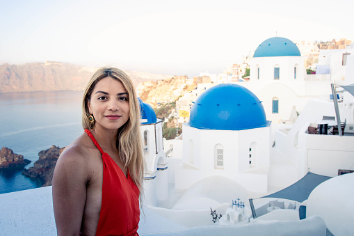 woman in red dress on vacation in Santorini Greece, a summer day. white houses with blue roofs are seen in the background.