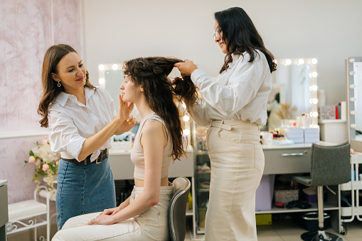 Side view of positive female make up artist and hairdresser prepare to doing makeup and hairstyle for young woman in beauty salon sitting in front of mirror. Four-handed makeup and styling concept.