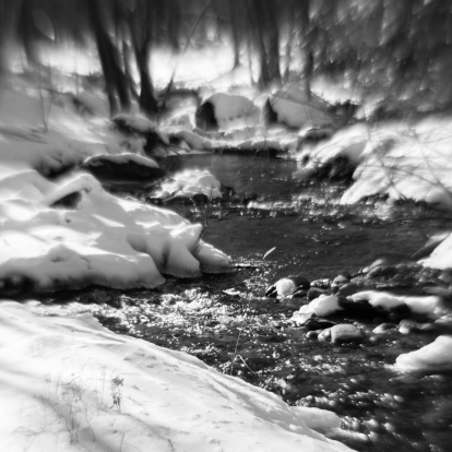 A black and white landscape photograph of Windows Creek in Pocono. The white of the riverbanks, which could be snow, contrasts against the darkness of the river. The outer edges of the image is blurred, while the middle is sharper and more focused.