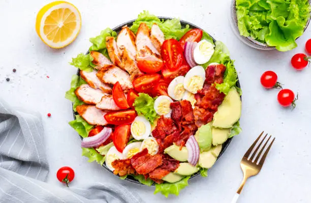 Photo of Traditional Cobb salad with chicken fillet, tomatoes, eggs, bacon, avocado and lettuce, gray table background, top view. American cuisine dish