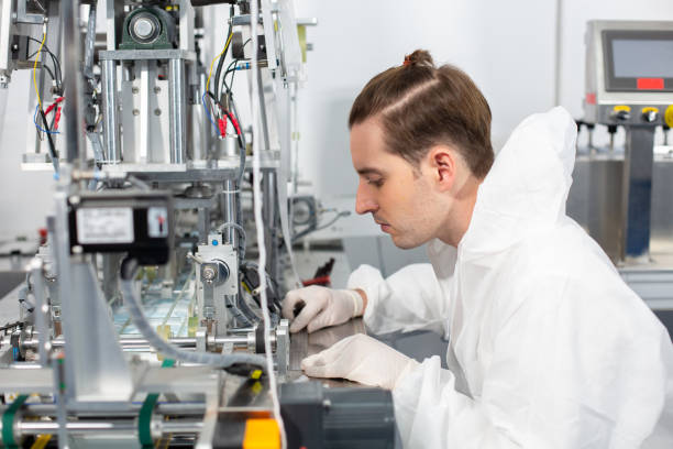 istock Machine staff, mask production department. Engineer wearing protective suit Inspecting quality of masks. machine production manufacturing factory. 1632749446