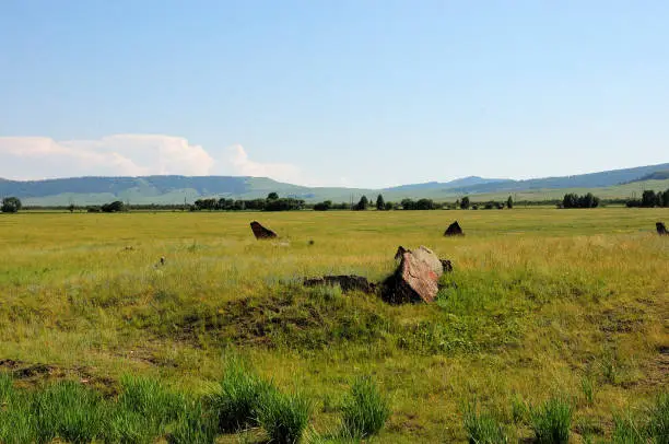 Photo of An ancient burial in the form of a low hill with a large burial stone standing in the middle of the endless steppe under a cloudy summer sky.