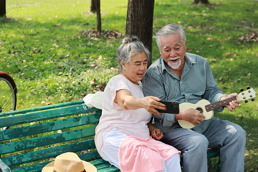 Happy smiling asian senior man and woman sitting on bench and using smartphone with ukulele in garden park outdoor. Musical and relaxation makes lover couple happiness. Health care lifestyle concept.