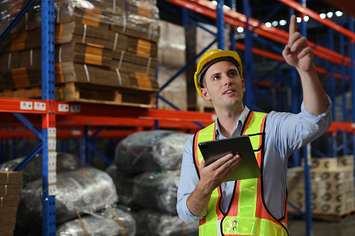 Warehouse workers man with hardhats and reflective jackets using tablet, walkie talkie radio and cardboard while controlling stock and inventory in retail warehouse logistics, distribution center