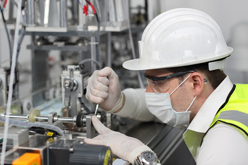 Caucasian mechanic technician maintenance, repairing industrial machinery equipment in factory. Professional worker in protective clothing with goggles and mask using wrench at manufacturing factory