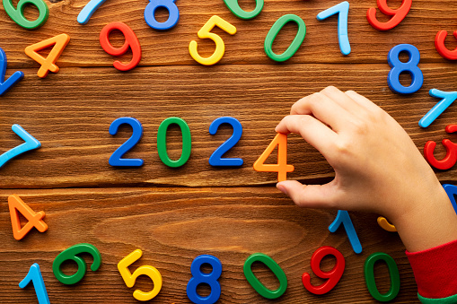 The child makes up the year 2024 from multi-colored numbers on a wooden background. New Year
