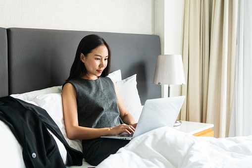 Businesswoman sitting on bed in hotel room while working on computer. Business trip, work from home concept. Copy space