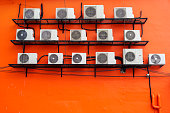 Air conditioners of various sizes are mounted on a bracket attached to the orange building wall