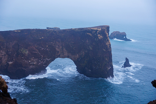 Stone coastline arch on dyrholaey peninsula with nordic foggy landscape and rocky cliffs. Spectacular shoreline stoney gate in iceland, beautiful wilderness with harbor bay view.