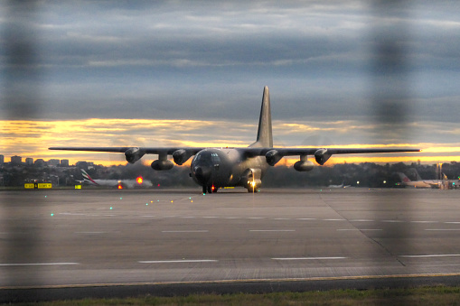 A Lockheed Martin C-130H plane, registration 61-PM, of the French Air Force taxiing before parking at Sydney Kingsford-Smith Airport. In the distance is an Emirates B777-31H(ER) plane, registration EK-EQF, having arrived from Dubai as flight EK416.  Other planes are visible on the right, parked at the international terminal.  This image was taken behind a steel security fence, causing the lines across the image.  This image faces west and was taken from Ross Smith Avenue, Mascot, on a cold, windy day after sunset on 20 August 2023.