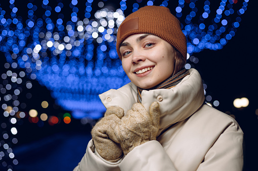 Winter portrait of female in warm clothes smiling and looking at camera against blurred background of bokeh glowing garlands in city at night
