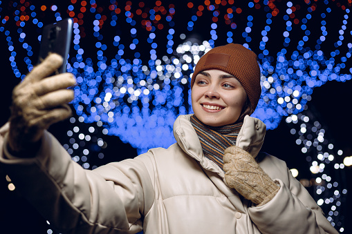 Positive female wearing warm hat gloves and jacket taking selfie on smart phone against bright festive glowing garlands in winter in city at night