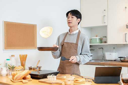 A joyful and handsome young Asian man in an apron, tossing pancakes on a frying pan, enjoys cooking pancakes in his kitchen.