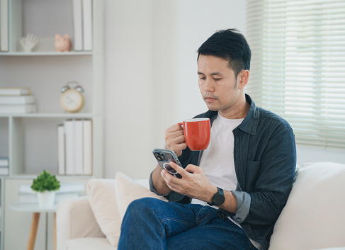 Asian man wearing casual clothes sitting on a couch sofa using mobile phone to social media or seach jobs or chatting or trade stock and drink hot of coffee at the living room.