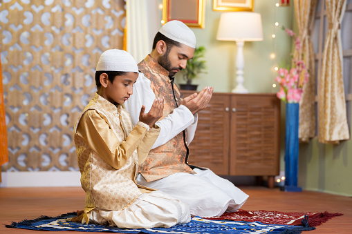Indian Muslim father with kid doing namaz or praying during Ramadan festival celebration at home - concept of Ramzan feast, traditions and bonding.