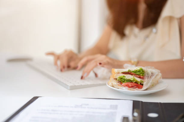 istock Close-up image of a sandwich on a table while a busy businesswoman works on a computer. 1633198880