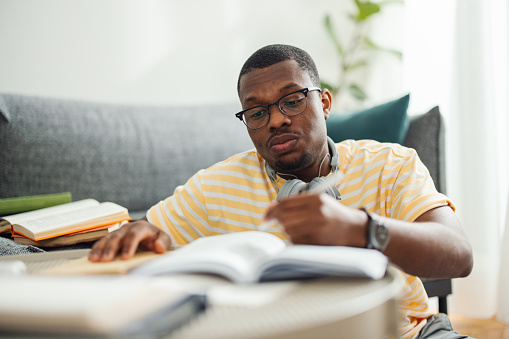 Close up shot of a serious African American man sitting on the floor with Bluetooth headphones around his neck, wearing glasses while writing notes in the notebook in front of him. He is studying for an exam or a test. There are several books around him.