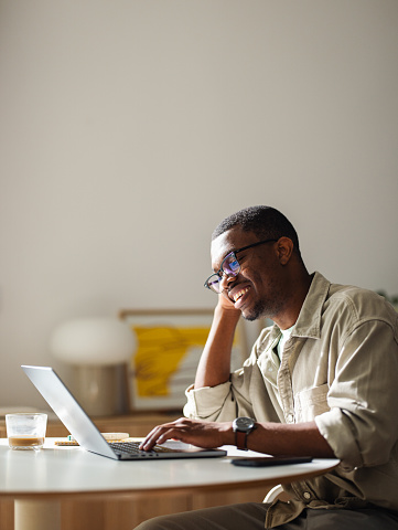 Close up shot of a smiling African American entrepreneur, wearing glasses, sitting at the table at home and using his laptop computer for remote work. He is looking down at the screen and smiling.