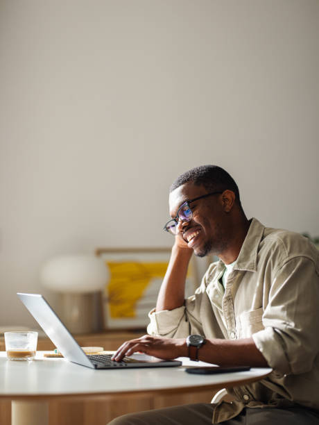 Portrait of an African American Entrepreneur Working From Home on His Laptop Computer Close up shot of a smiling African American entrepreneur, wearing glasses, sitting at the table at home and using his laptop computer for remote work. He is looking down at the screen and smiling. black man laptop stock pictures, royalty-free photos & images