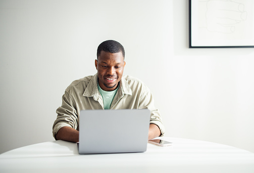 Close up shot of a handsome African American man sitting at the table and using his laptop computer for work. He is looking down at the screen and smiling.