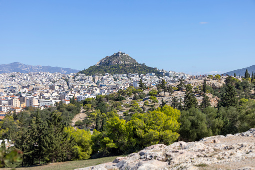 View of Athens, Greece, under the brilliance of a sunny summer day, with the iconic Mount Lycabettus towering over downtown. From this vantage point, the city sprawls out beneath the hill, showcasing a blend of ancient and modern architecture. Lycabettus Hill stands as a timeless sentinel, offering a stunning backdrop to the vibrant urban landscape. As the sunlight bathes the scene in warmth, immerse yourself in the beauty of Athens and the commanding presence of Mount Lycabettus, capturing the essence of a perfect summer day in the Greek capital.
